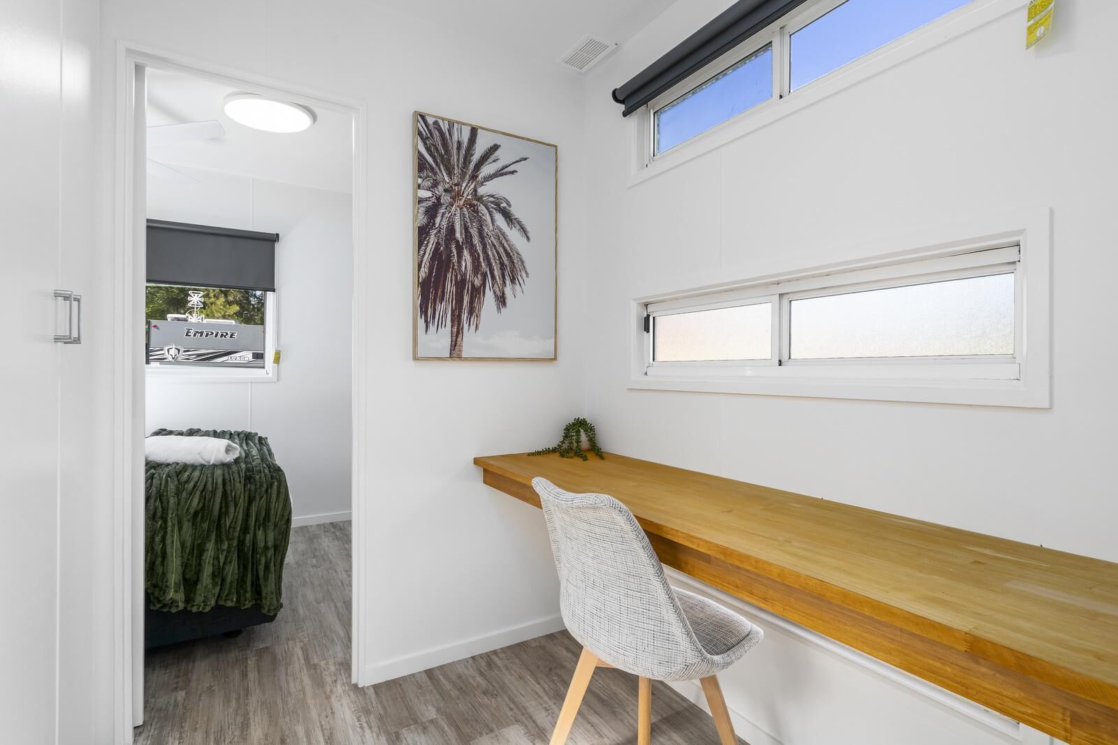 Ein moderner, hell eingerichteter Raum mit Schreibtisch aus Holz, einem Sessel, einem gerahmten Bild und einem Fenster mit Jalousien.