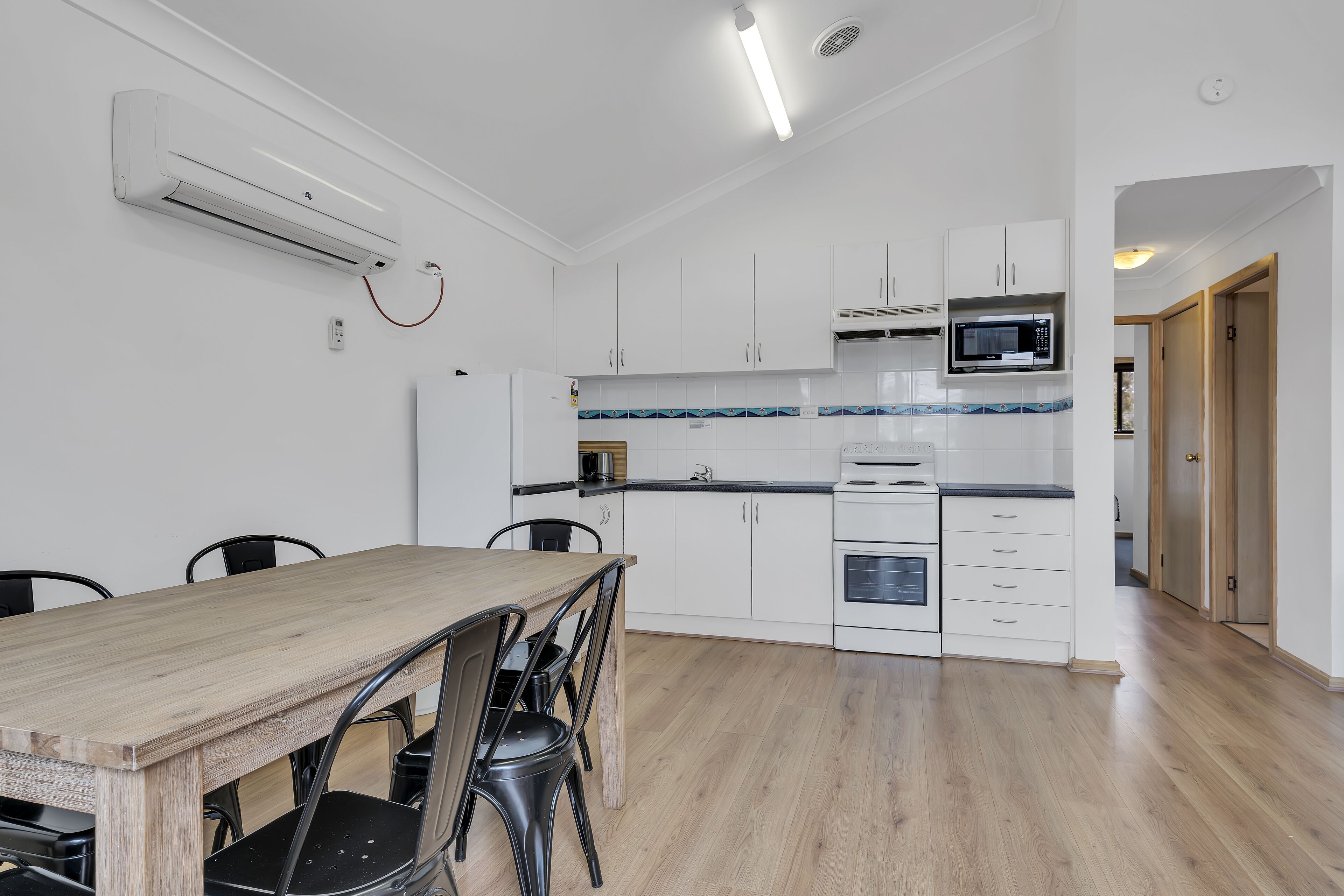 A modern kitchen with white cabinets, a microwave, and a built-in oven. There is a dining table with chairs and a ceiling-mounted air conditioner.