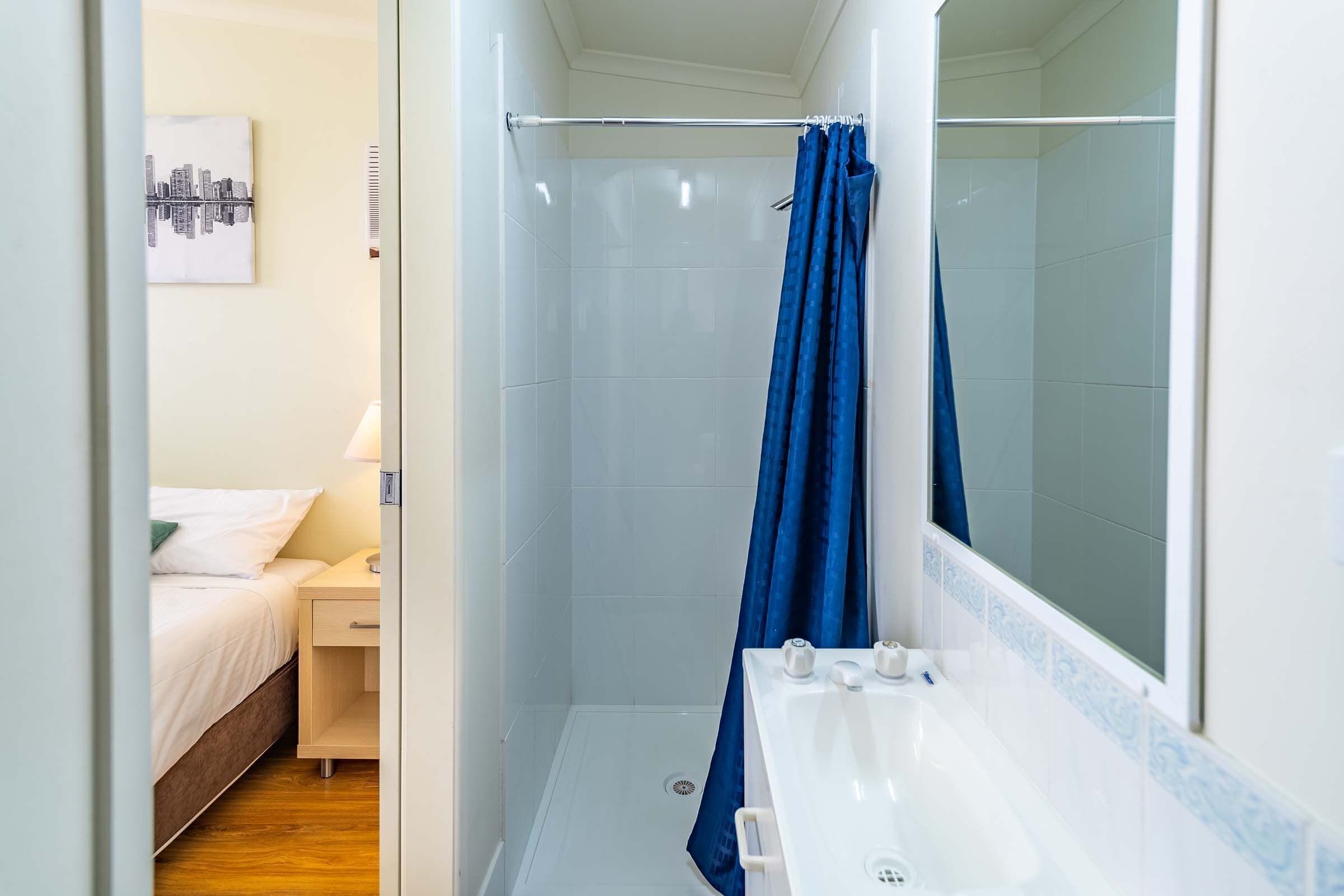 Un baño pequeño con ducha, lavamanos y una cama visible en la habitación contigua.