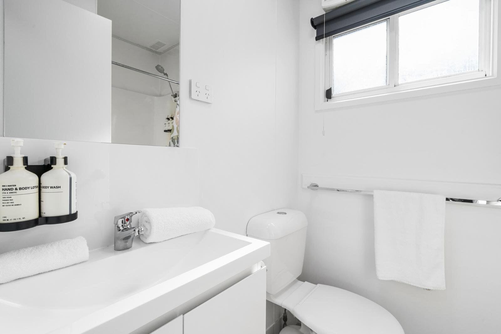 A modern bathroom with a white sink, a toilet, a towel rack, and a window with a blind.