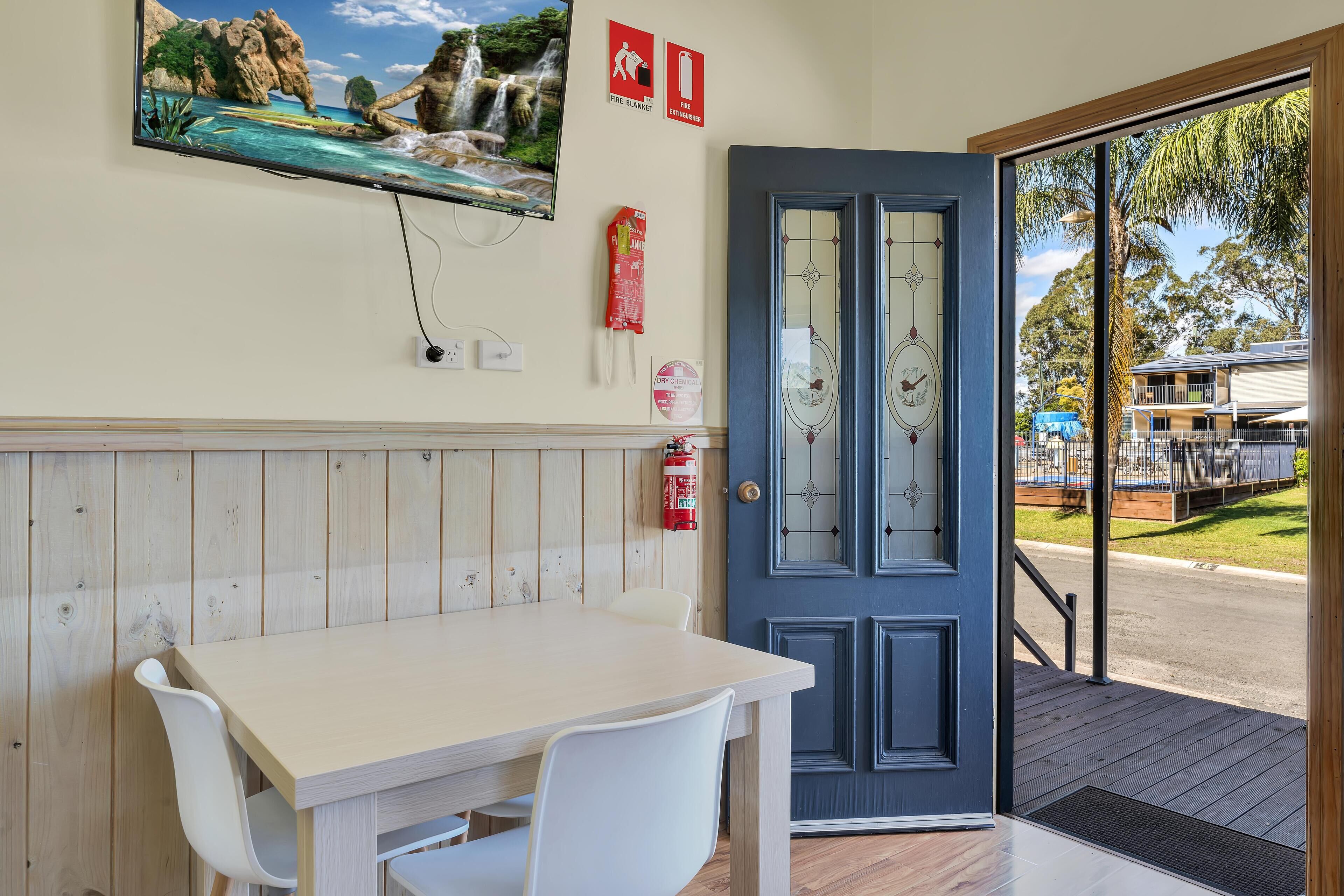 A room with a wooden panel wall, a white table with chairs, a fire extinguisher, and a door leading outside.
