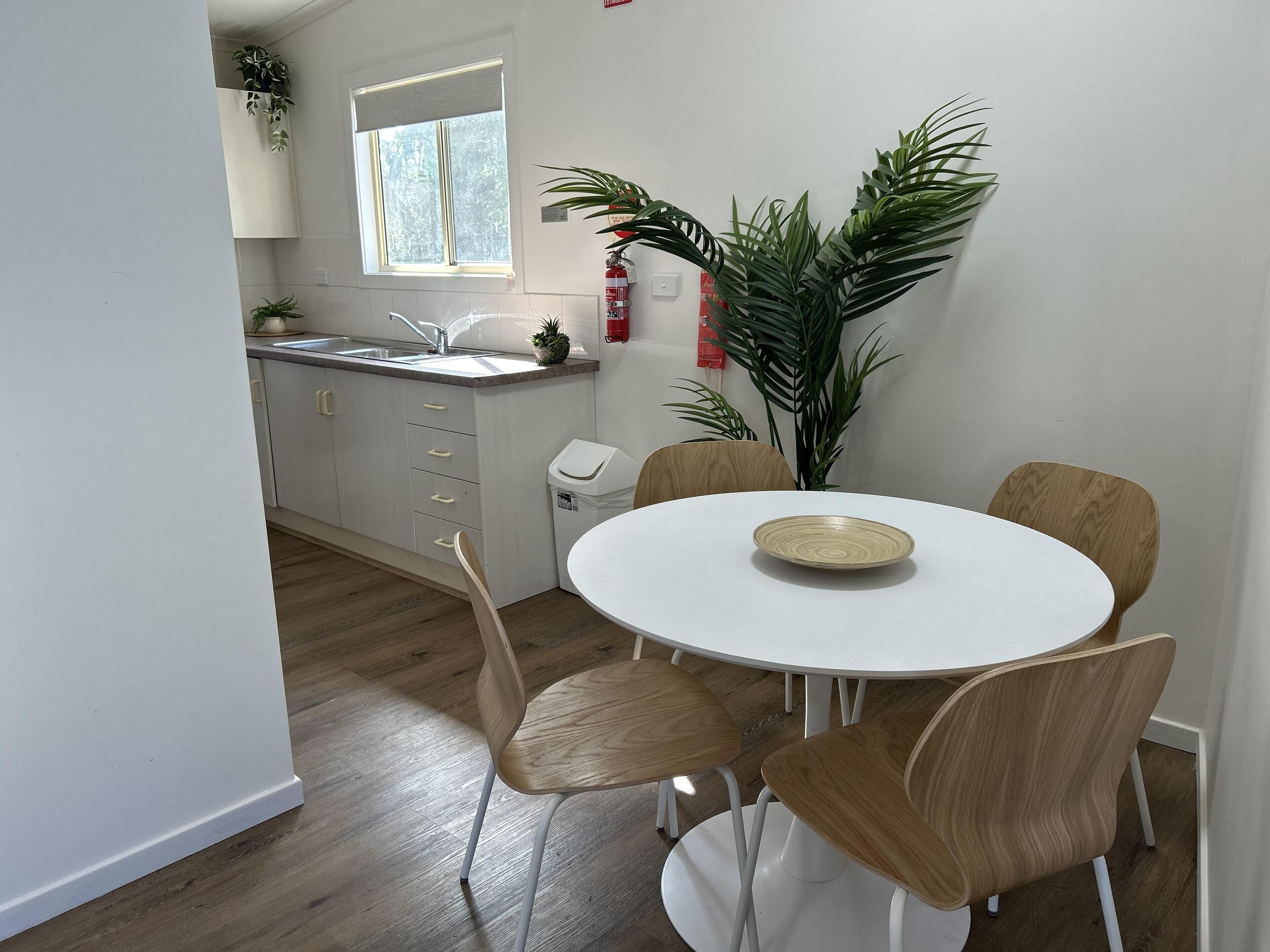 Une cuisine moderne avec des meubles blancs, un évier et une fenêtre. On y trouve une table à manger avec quatre chaises et une assiette. Une grande plante se trouve dans un coin.