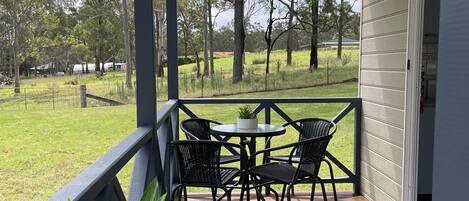 A covered outdoor area with a wooden deck, a table with chairs, and a view of a grassy field and trees.