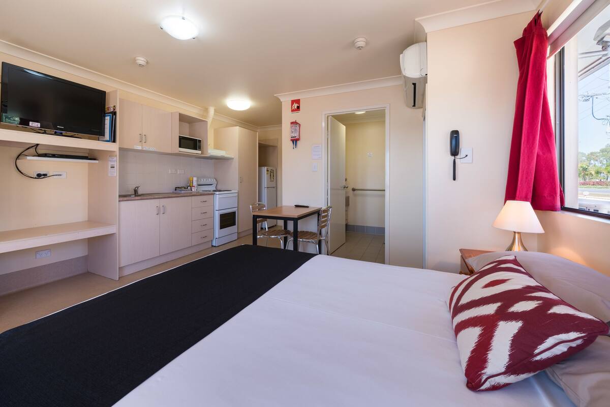 Habitación de hotel compacta con cama, escritorio, kitchenette y televisor.