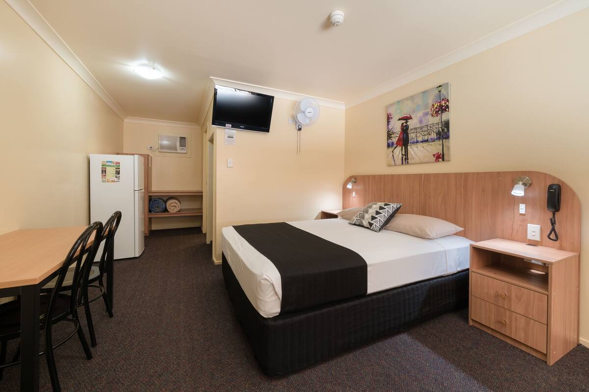 Habitación de hotel con cama, escritorio, silla, refrigerador, televisión, un cuadro y un teléfono en la mesita de noche.