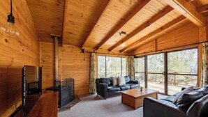 A wooden cabin interior with a fireplace, leather sofas, a coffee table, and large windows offering a view of the outdoors. - Curringa Farm (Hamilton)