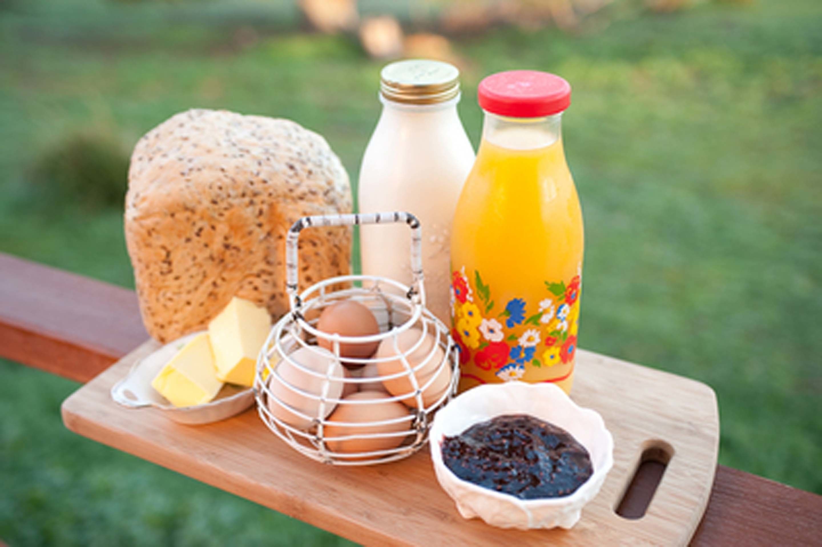 Una mesa de madera con un queso redondo, una botella de leche, una botella de jugo, una cesta de alambre con huevos, un plato con mantequilla y un recipiente con mermelada.