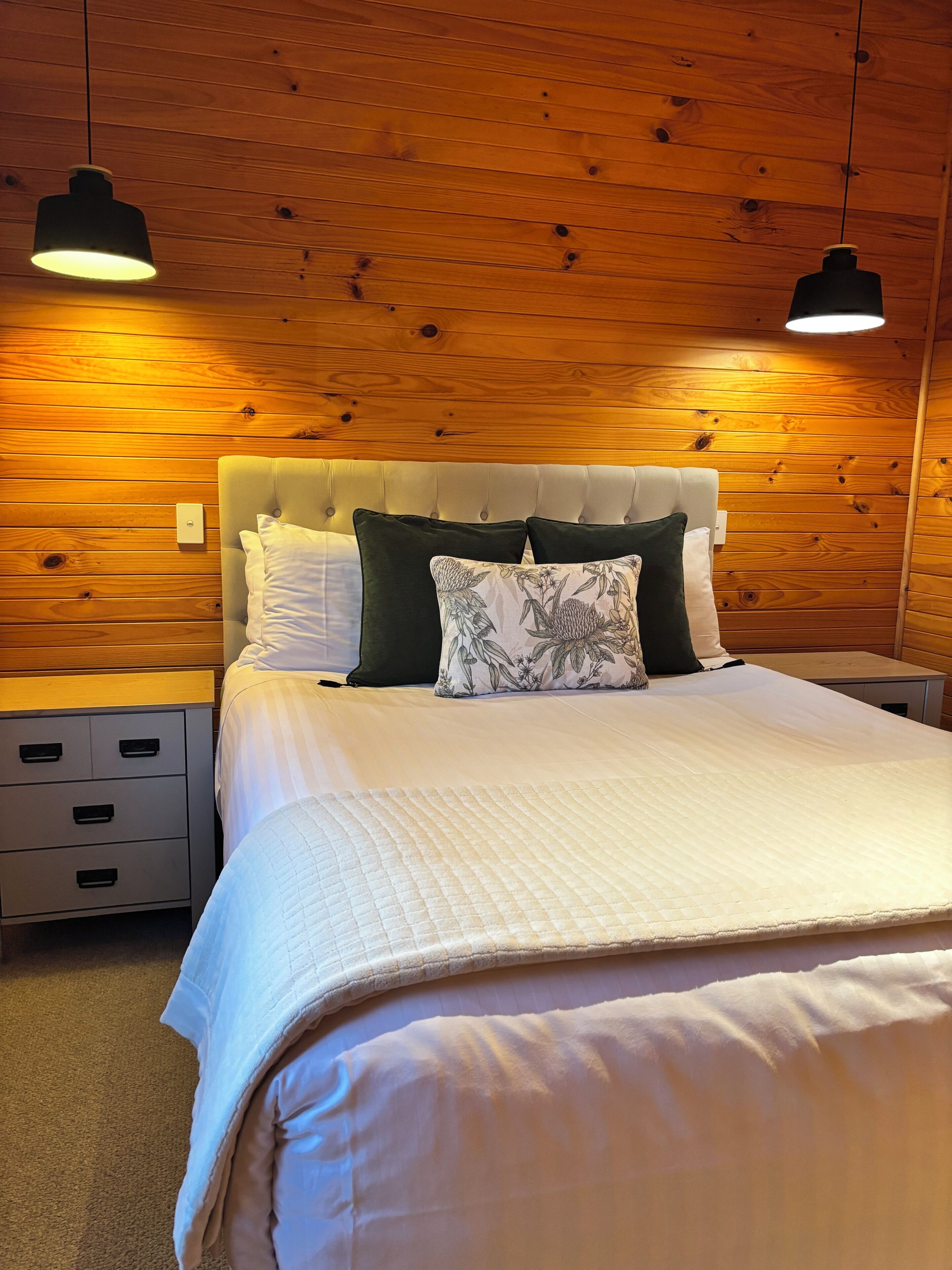 A neatly made bed with a tufted headboard, a bedside table with drawers, and two pendant lights.
