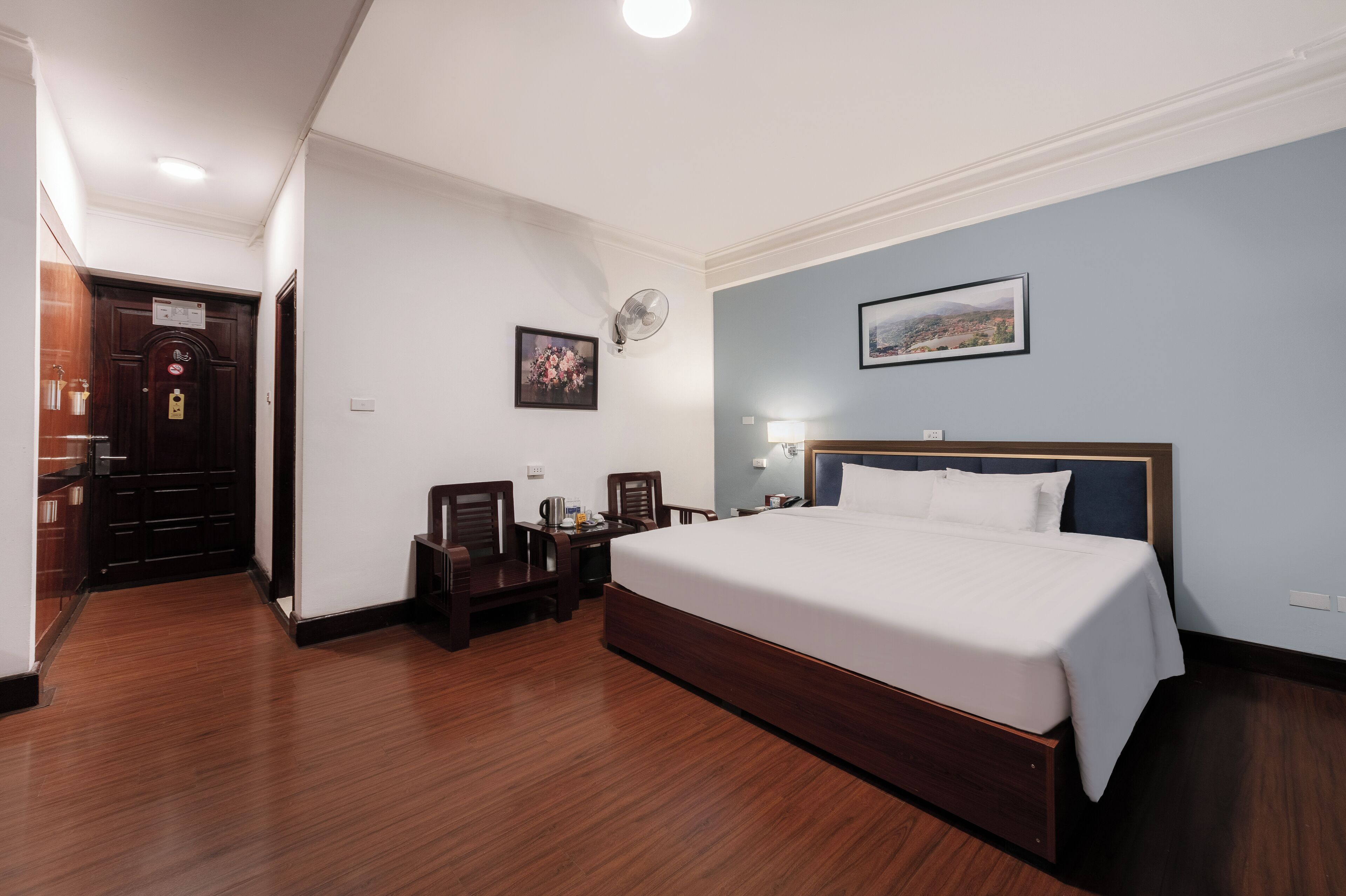A hotel room with a large bed, a desk with a chair, a small table with a lamp, and a picture on the wall.