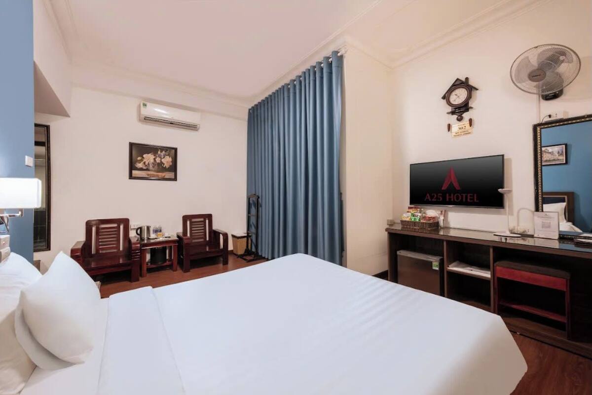 Habitación de hotel con cama, escritorio, dos sillas, televisor, reloj y ventilador.