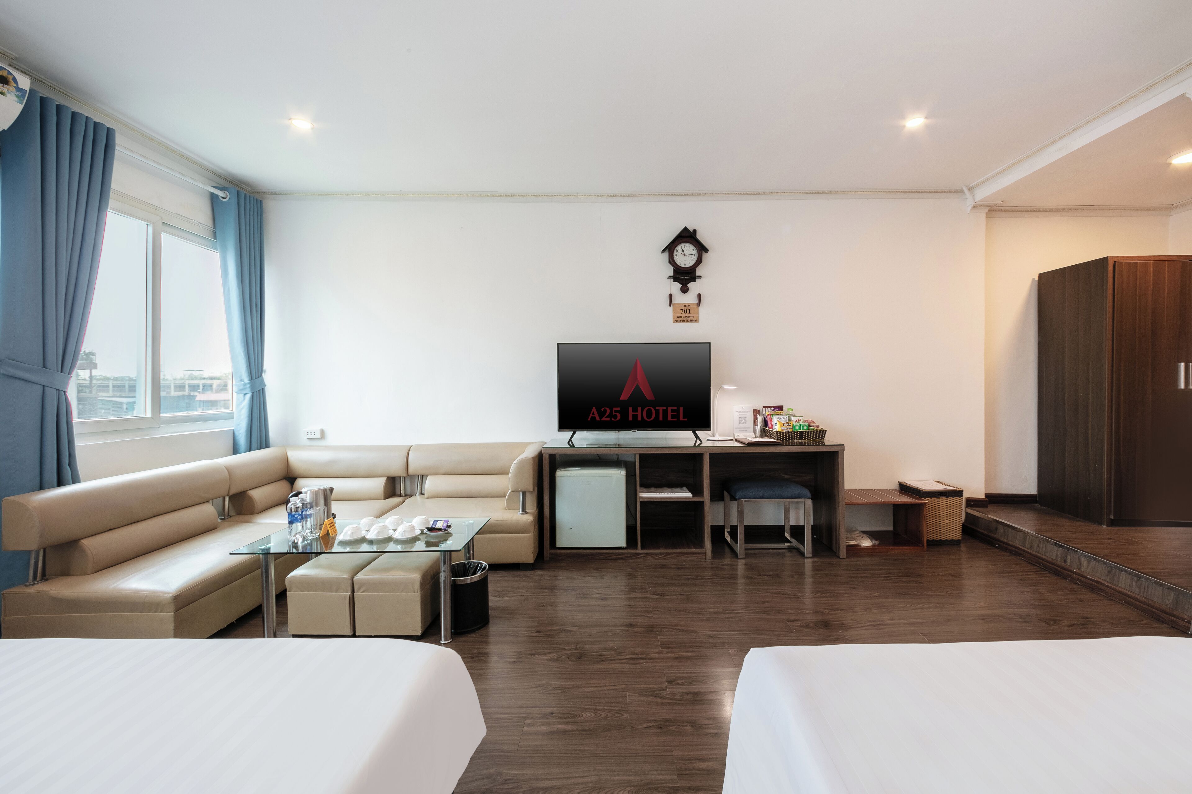 A hotel room with a sofa, two beds, a desk with a TV, a small fridge, and a window with curtains.