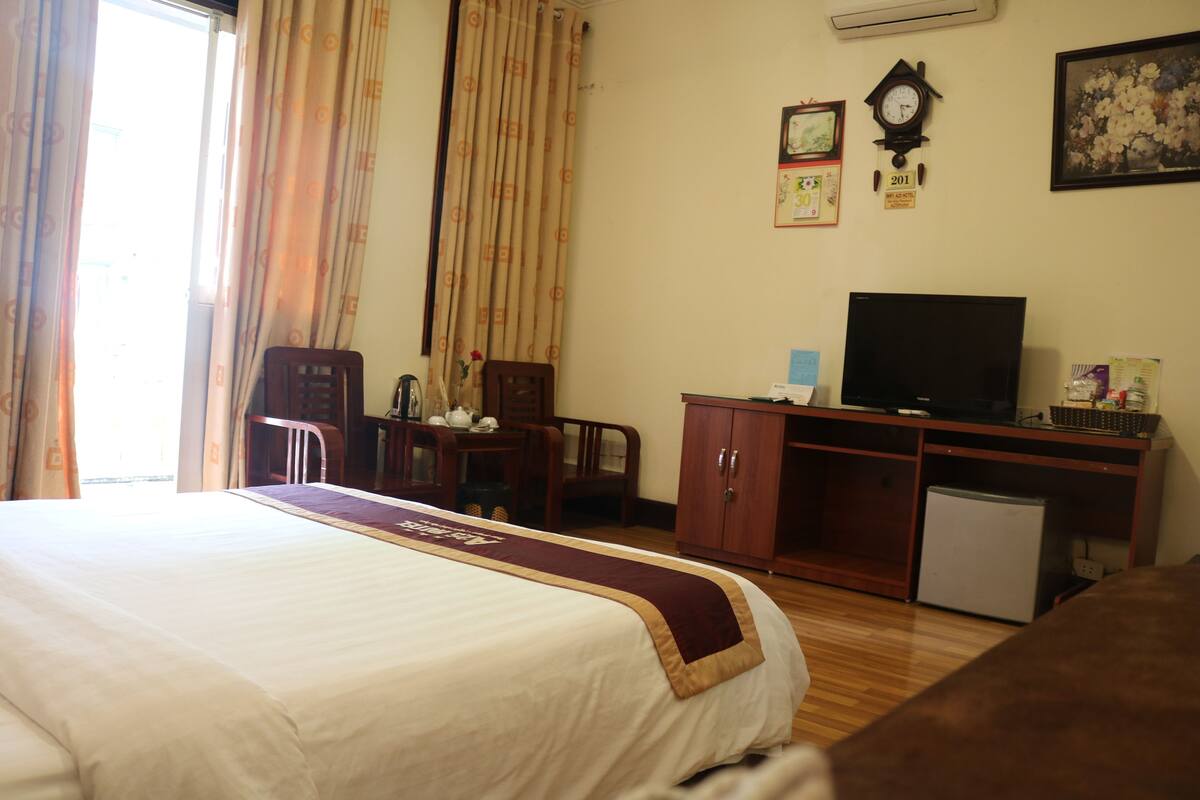 Habitación de hotel con cama, escritorio con televisor, refrigerador pequeño y un reloj en la pared.