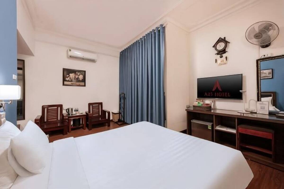 Una habitación de hotel con cama, dos sillas, un escritorio con computadora, televisor y ventilador.