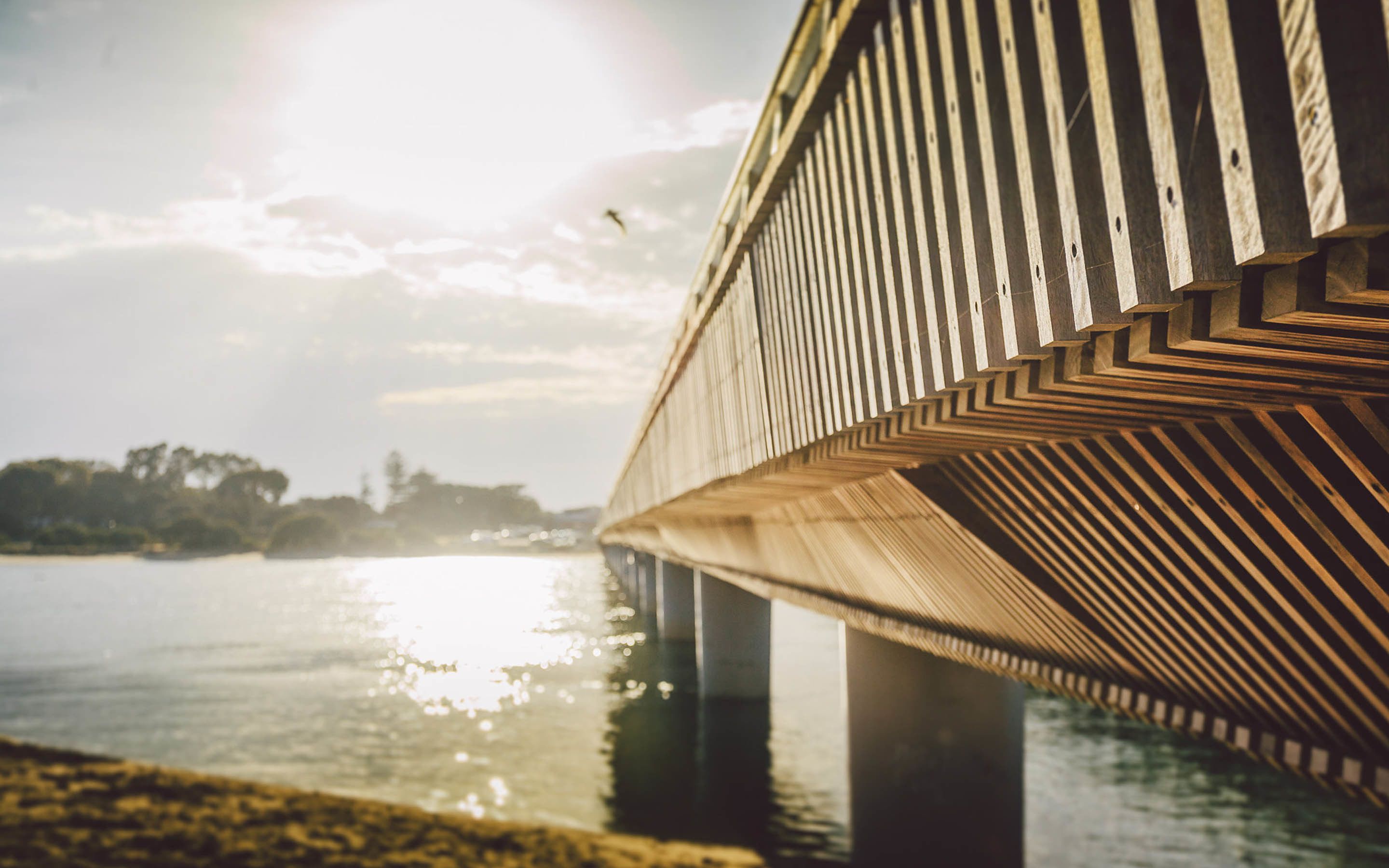 A wooden bridge with a chevron pattern, over a body of water, with a clear sky and sunlight reflecting on the surface.