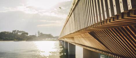 Un pont en bois orné d’un motif en chevrons, enjambant une étendue d’eau, avec un ciel dégagé et la lumière du soleil se reflétant à la surface.