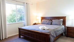 A neatly made bed with a wooden headboard, a nightstand with a lamp, and a window with curtains. - Point Lonsdale Holiday Apartments - Adults only (Point Lonsdale)