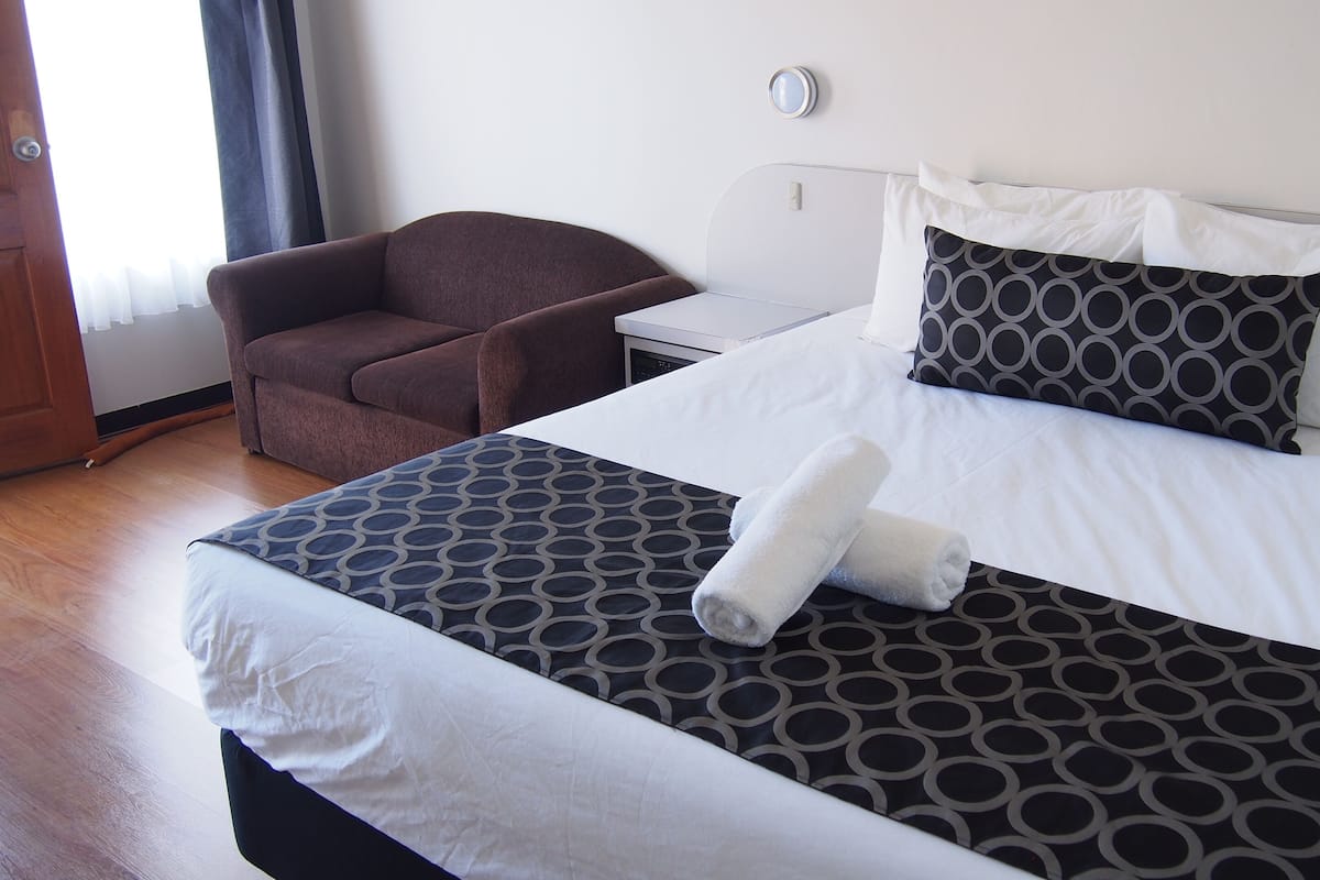 Habitación de hotel con una cama grande, un sillón marrón, una mesita de noche y un espejo en la pared.