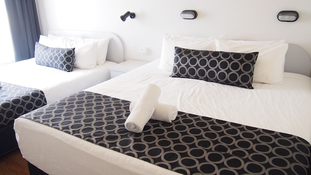 Habitación de hotel con dos camas, cada una con colcha y almohadas estampadas, una mesita de noche blanca y una lámpara de pared.