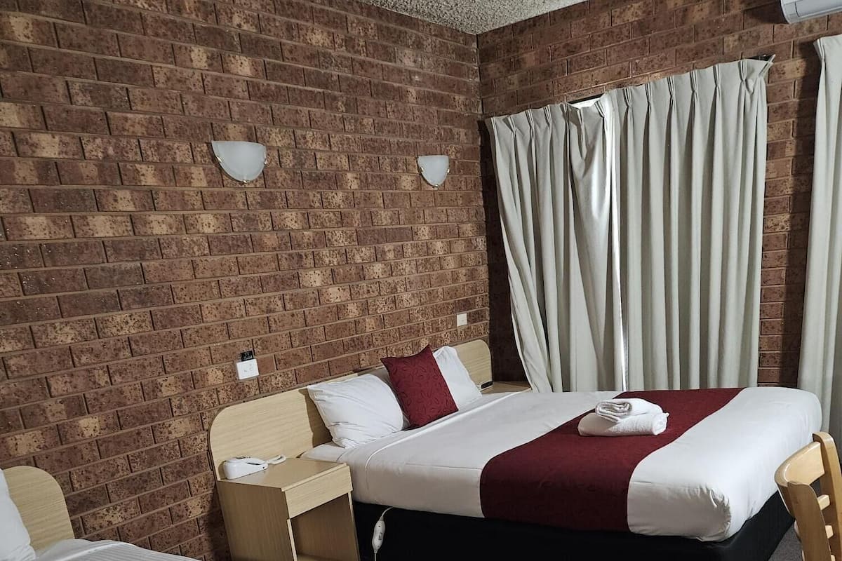 A hotel room with two beds, a brick wall, a window with curtains, and a small table with a lamp.