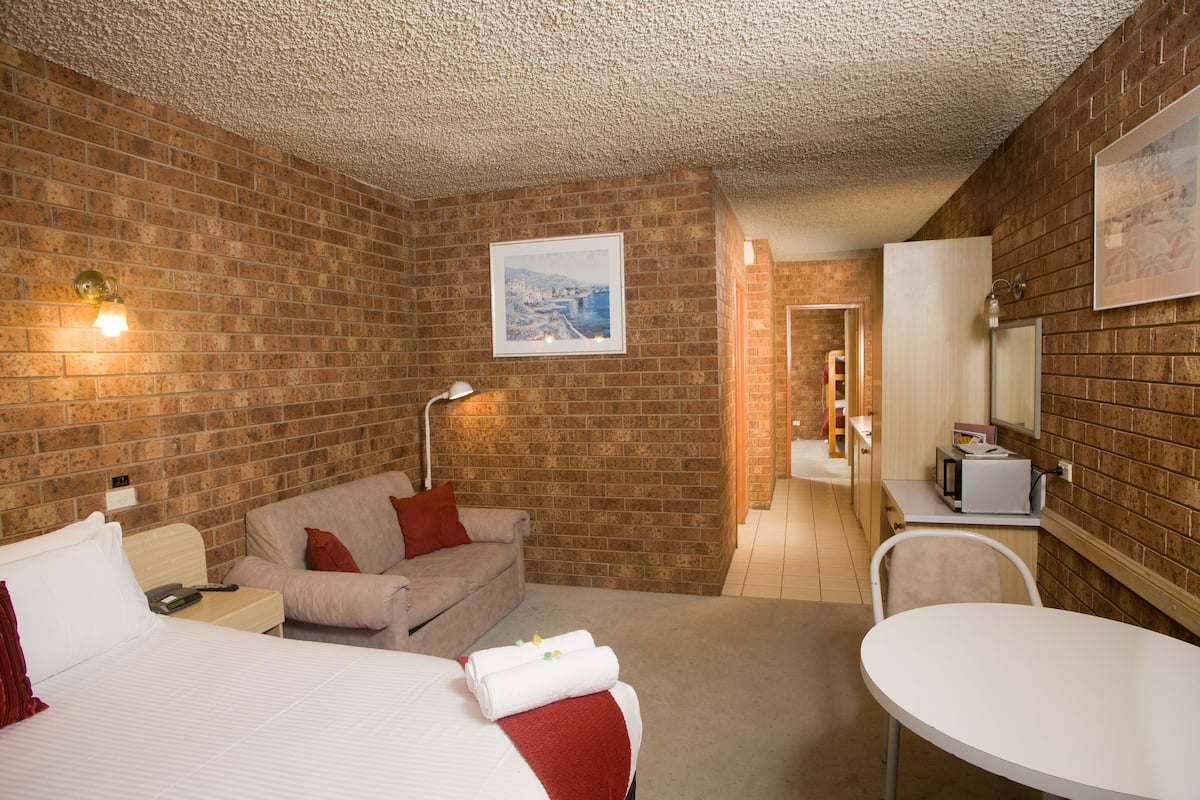 A hotel room with a brick wall, a bed, a sofa, a dining table, and a microwave.
