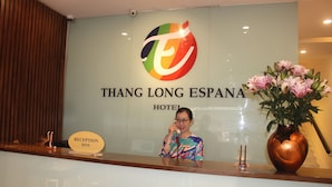 A reception desk at Thang Long Espana Hotel with a logo, a reception sign, and a vase of flowers.