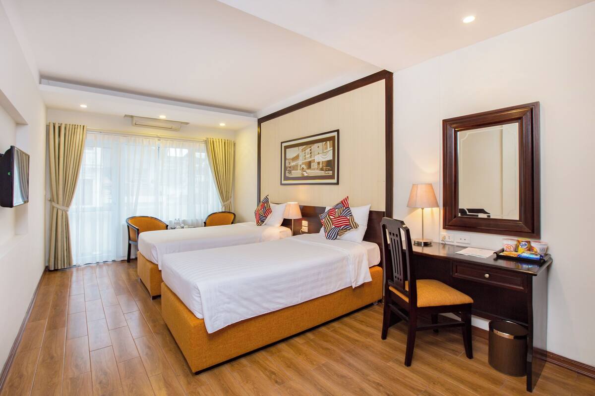 Une chambre d’hôtel avec deux lits, un bureau avec une chaise, un miroir et une fenêtre avec des rideaux.