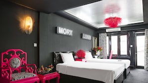 A modern hotel room with two beds, a bright ceiling light, and a large window with patterned curtains.