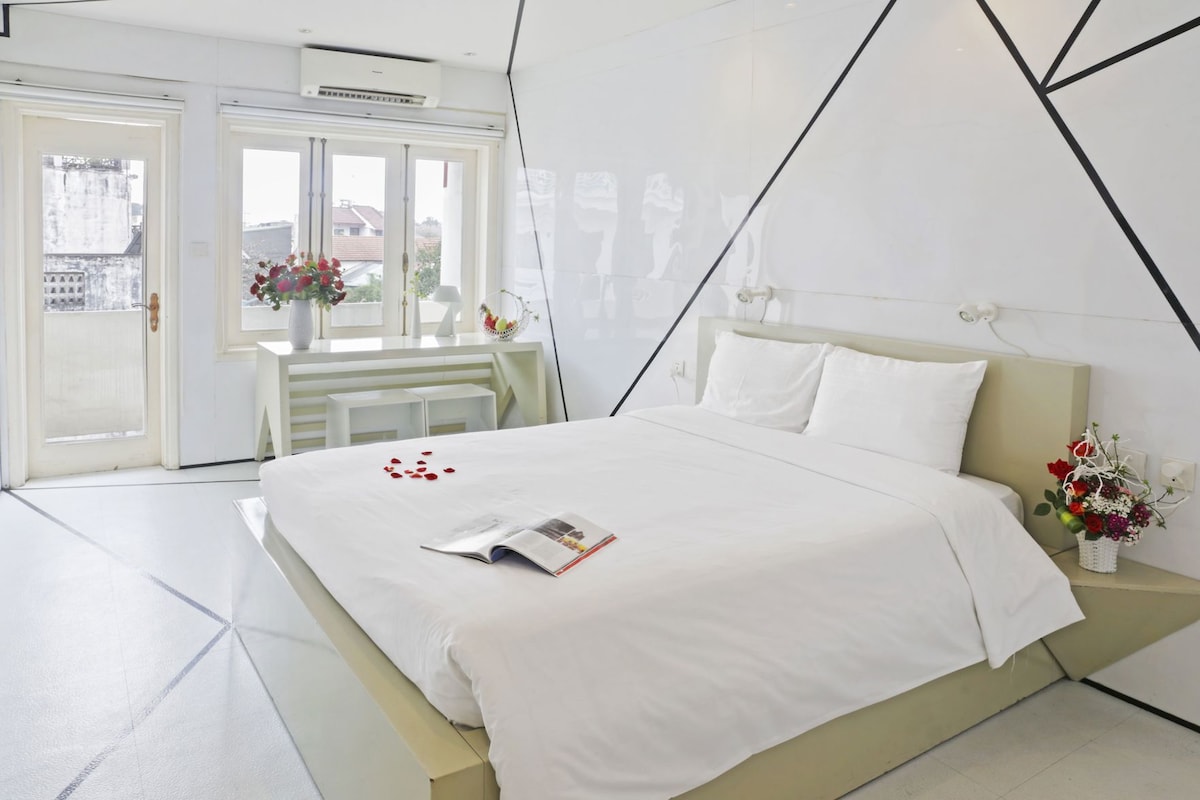 Une chambre d’hôtel moderne, équipée d’un lit, d’une table de chevet agrémentée d’un vase de fleurs et d’une fenêtre donnant sur des immeubles.
