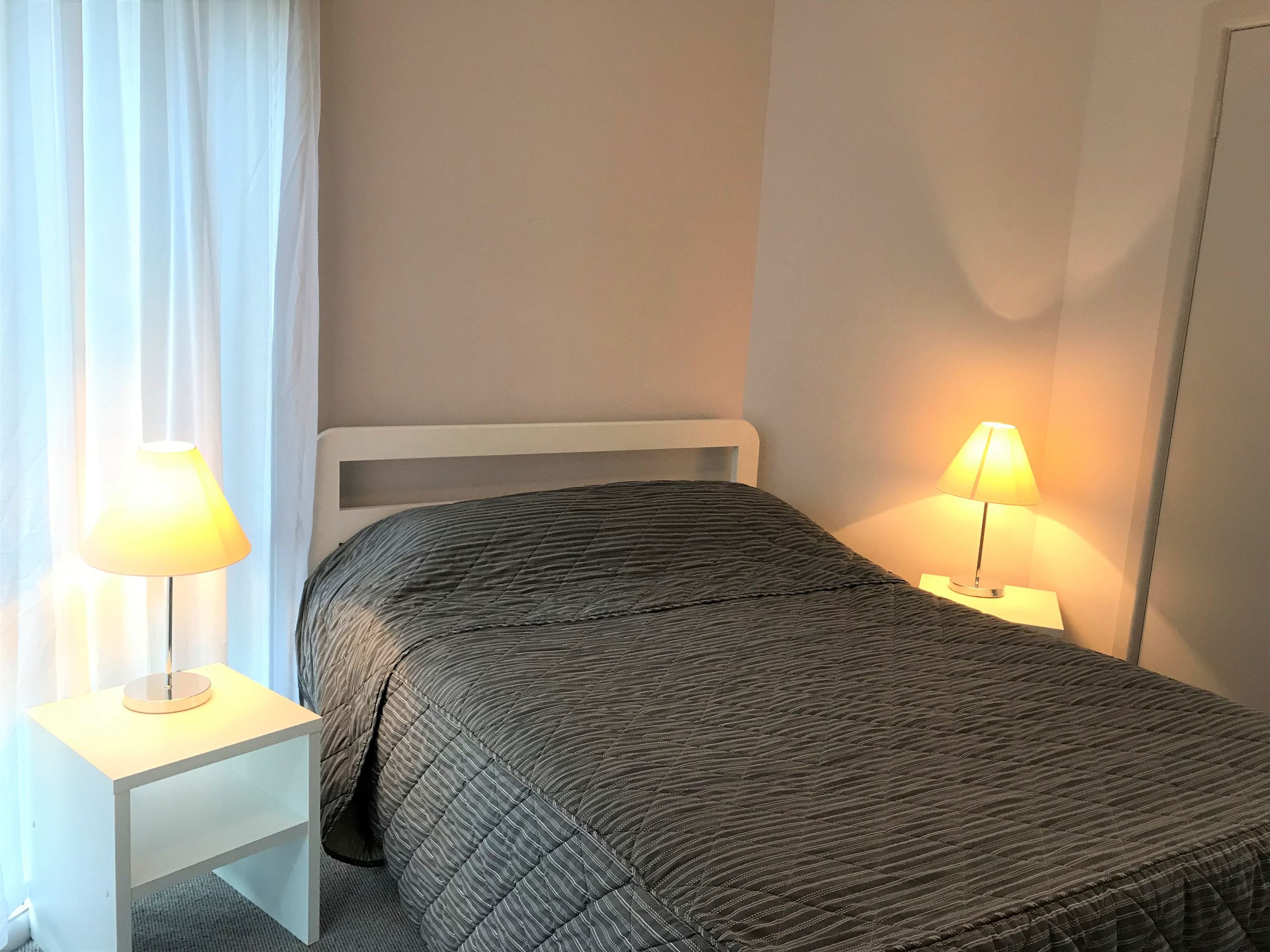 A neatly made bed with a grey quilt, two bedside lamps, and a white nightstand.