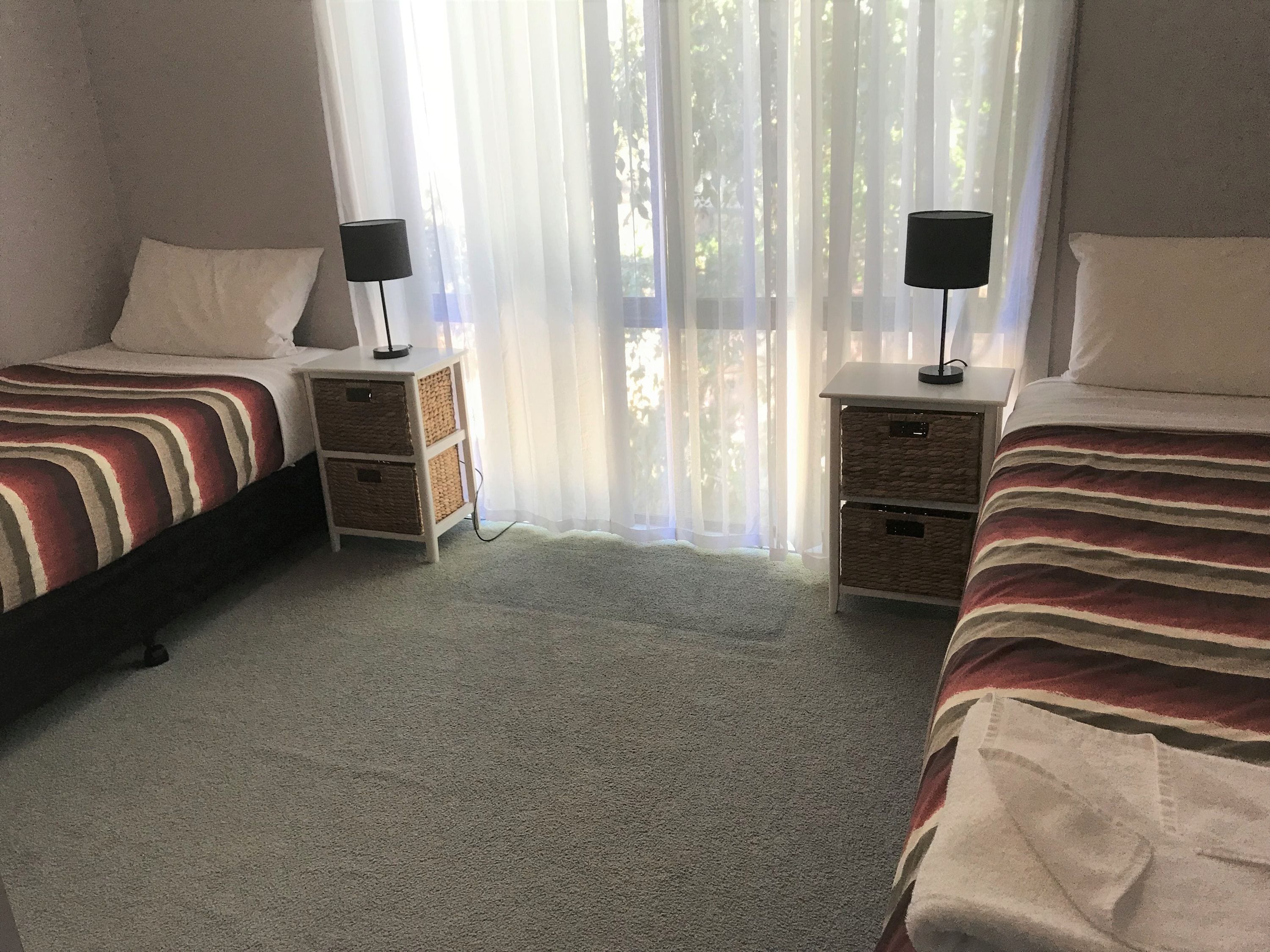 A bedroom with two beds, each with a bedside table and lamp, and a window with sheer curtains.