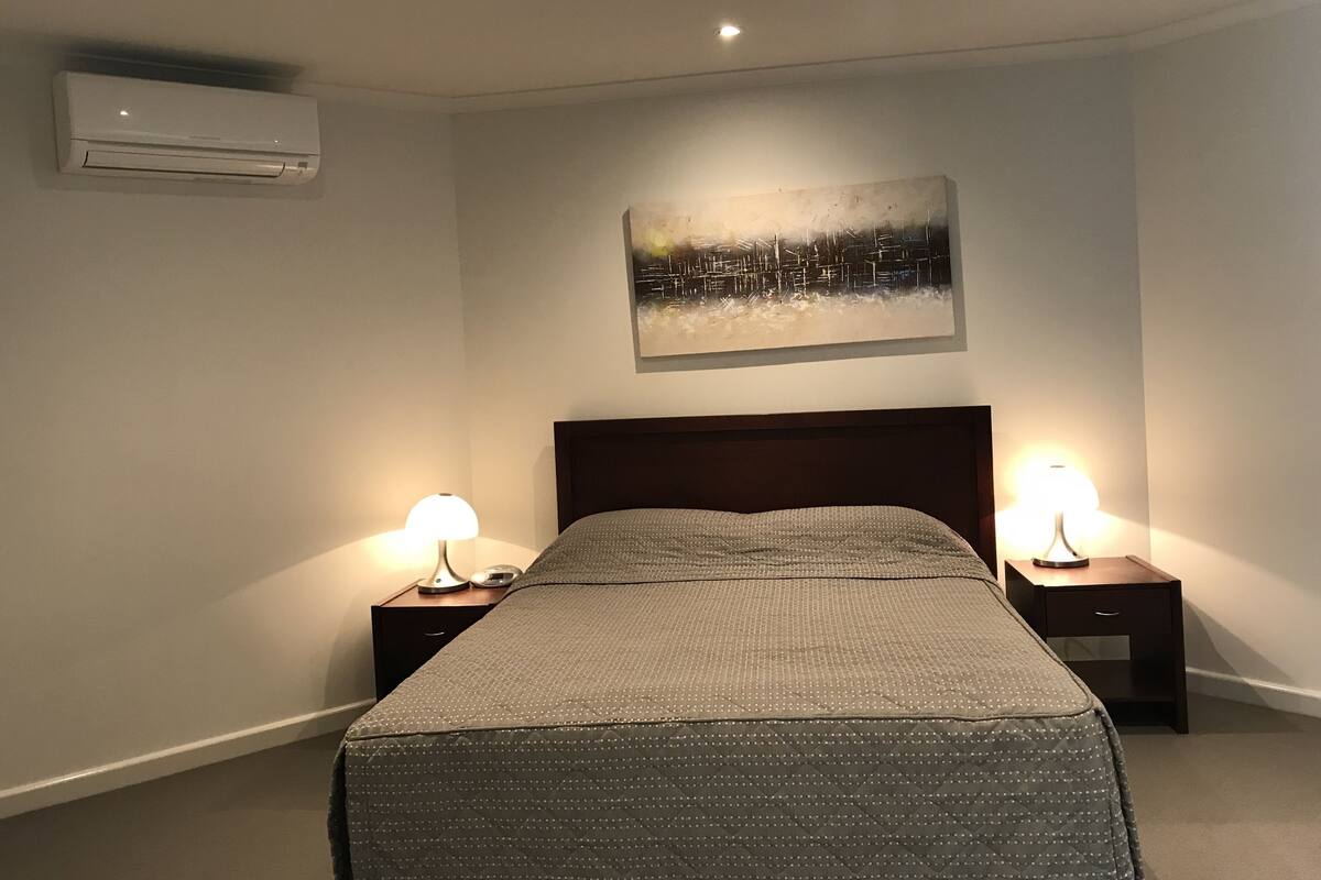 A hotel room with a bed, two bedside tables with lamps, an air conditioner, and a cityscape painting on the wall.