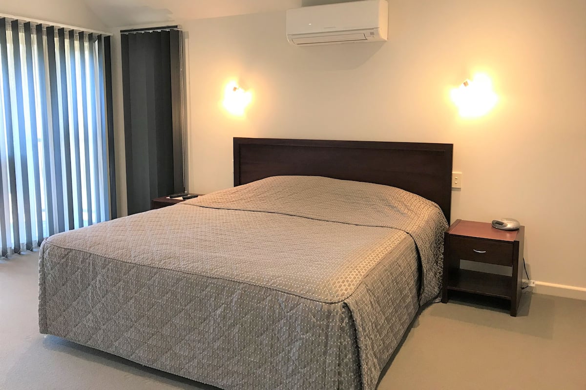 A neatly made bed with a quilted cover, a bedside table with a lamp, and an air conditioning unit mounted on the wall.