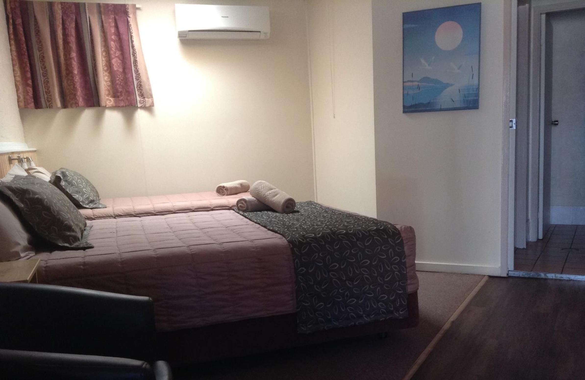 Un dormitorio con dos camas, una mesita de noche, una lámpara, un cuadro y una puerta que da a otra habitación.