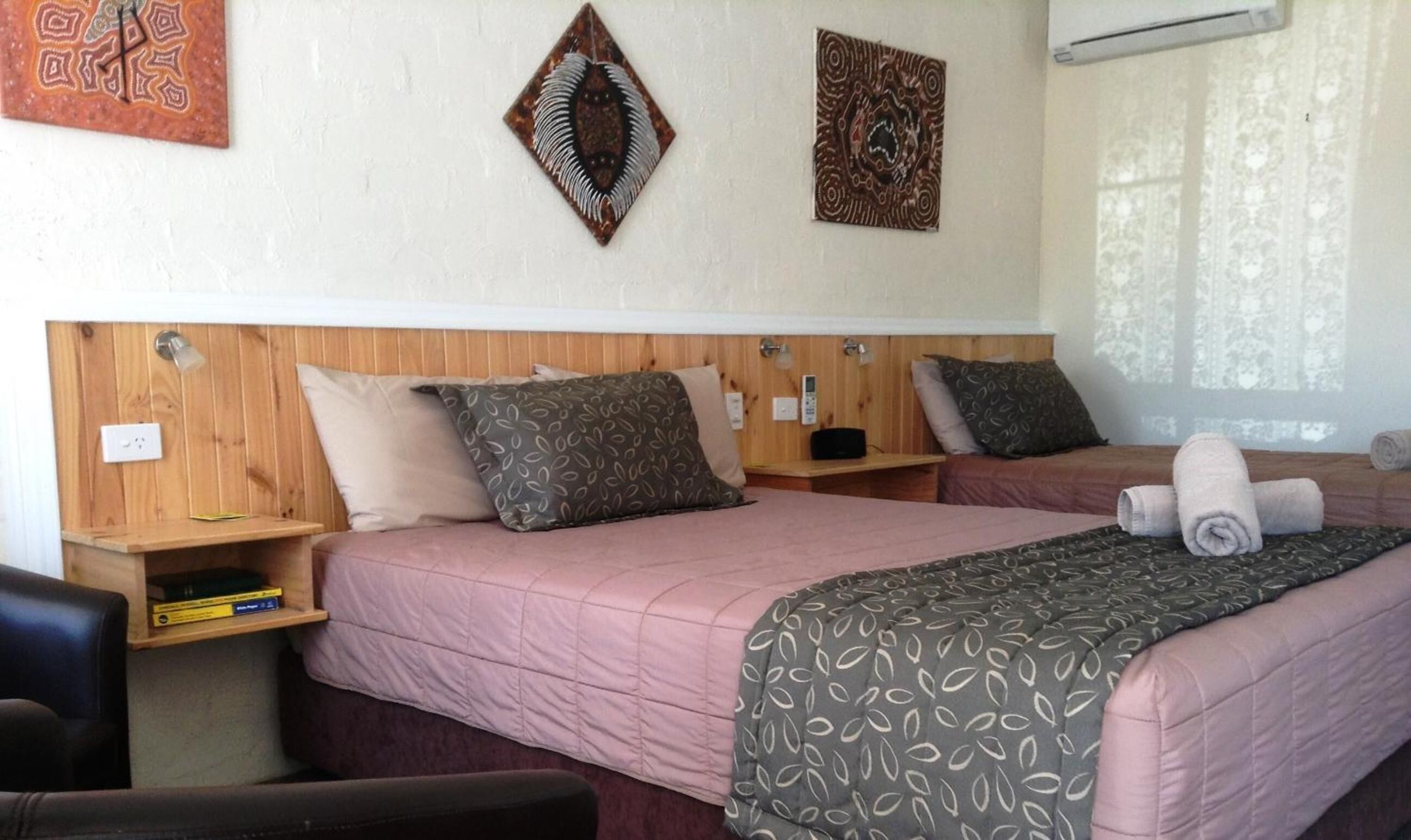 Una habitación de hotel con dos camas, cabeceros de madera y una ventana con cortinas translúcidas.