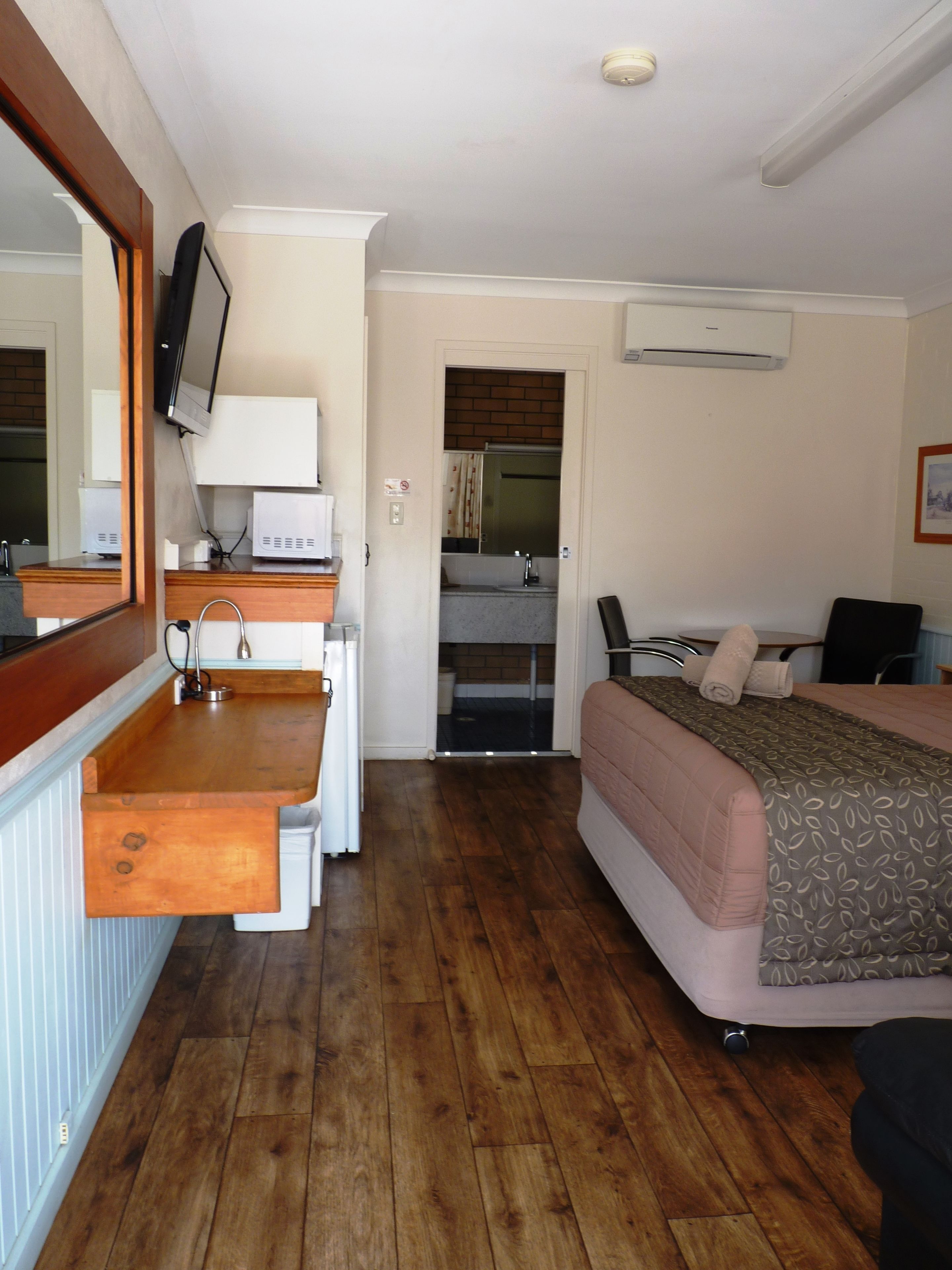 Een compacte kamer met een houten vloer, een bed met een geborduurde deken, een bureau met een computer, een spiegel en een aan de muur bevestigde televisie.