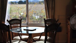Un coin repas avec des chaises en osier et une table en verre, une fenêtre donnant sur un champ et des montagnes, et un ventilateur sur le côté droit.
