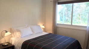Una cama bien hecha con una colcha escocesa, dos lámparas de noche y una ventana con vistas a árboles.