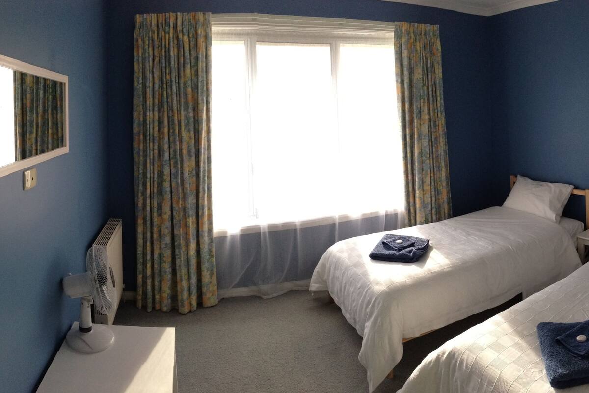 Een hotelkamer met twee bedden, een raam met gordijnen, een radiator en een lijst met een afbeelding aan de muur.