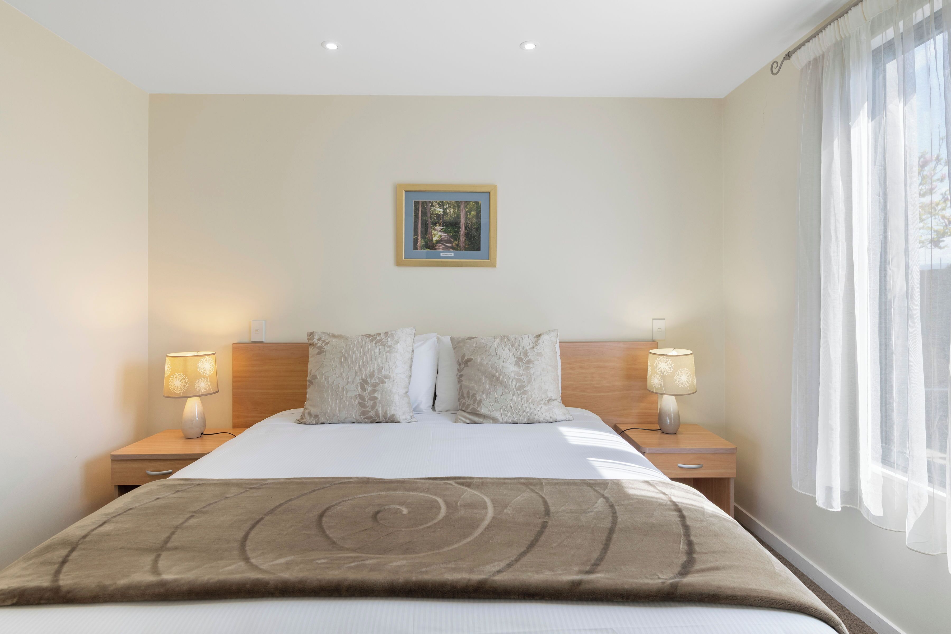 Ein Hotelzimmer mit einem Bett, zwei Nachttischlampen, einem gerahmten Bild an der Wand und einem Fenster mit durchscheinenden Vorhängen.