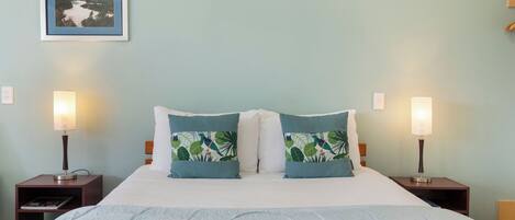 A neatly made bed with a blue blanket, two pillows with a tropical leaf pattern, and two bedside lamps on wooden nightstands.