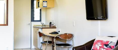Ett kompakt bostadsutrymme med ett litet kök, en matplats med två stolar, en soffa med en dekorativ kudde och en väggmonterad tv.
