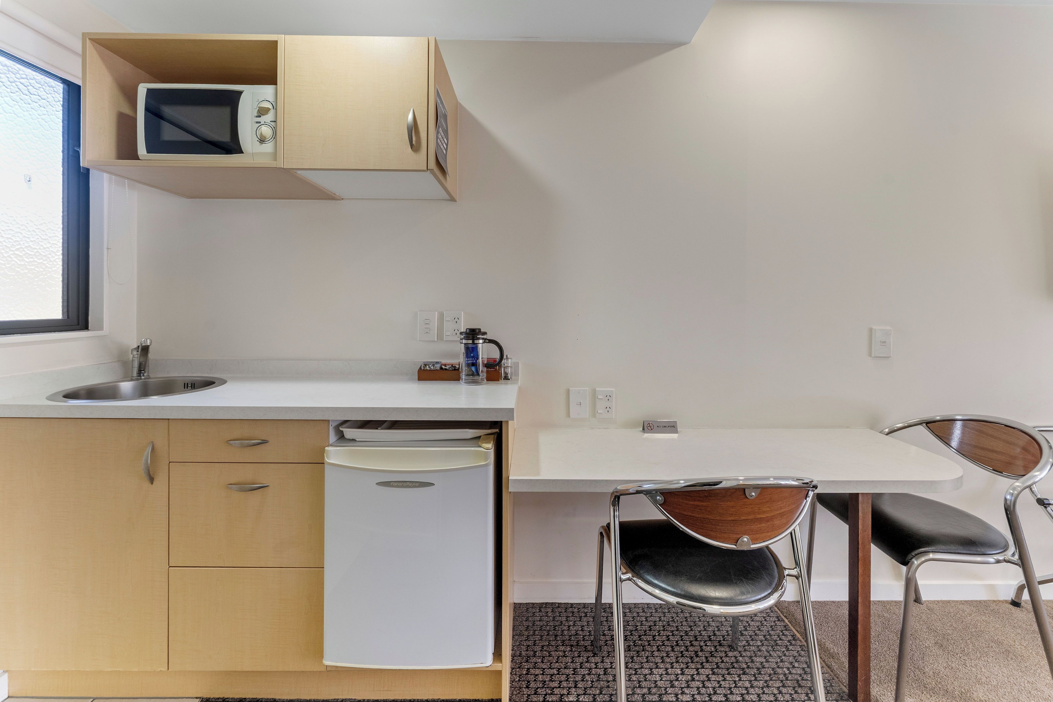 A compact kitchenette with a microwave, a sink, and a dishwasher. There is a small table with two chairs, a window, and a 'No Smoking' sign on the wall.
