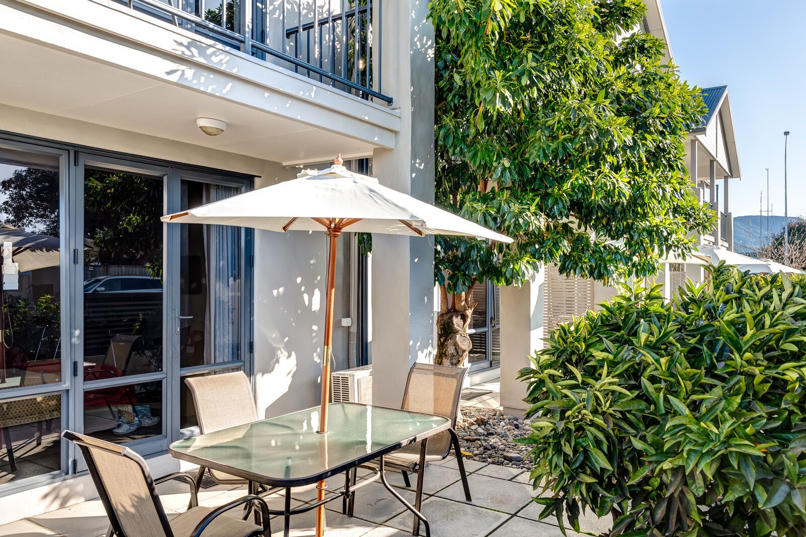 Une terrasse extérieure avec une table et des chaises en verre, un parasol et une vue sur un immeuble moderne doté de grandes fenêtres et d’un balcon.