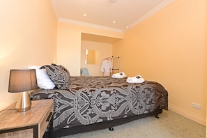 Habitación de hotel con una cama grande, una mesita de noche con lámpara, un espejo y un perchero con bata y zapatos.