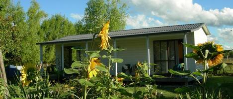 A single-story house with a porch, surrounded by lush greenery and sunflowers.