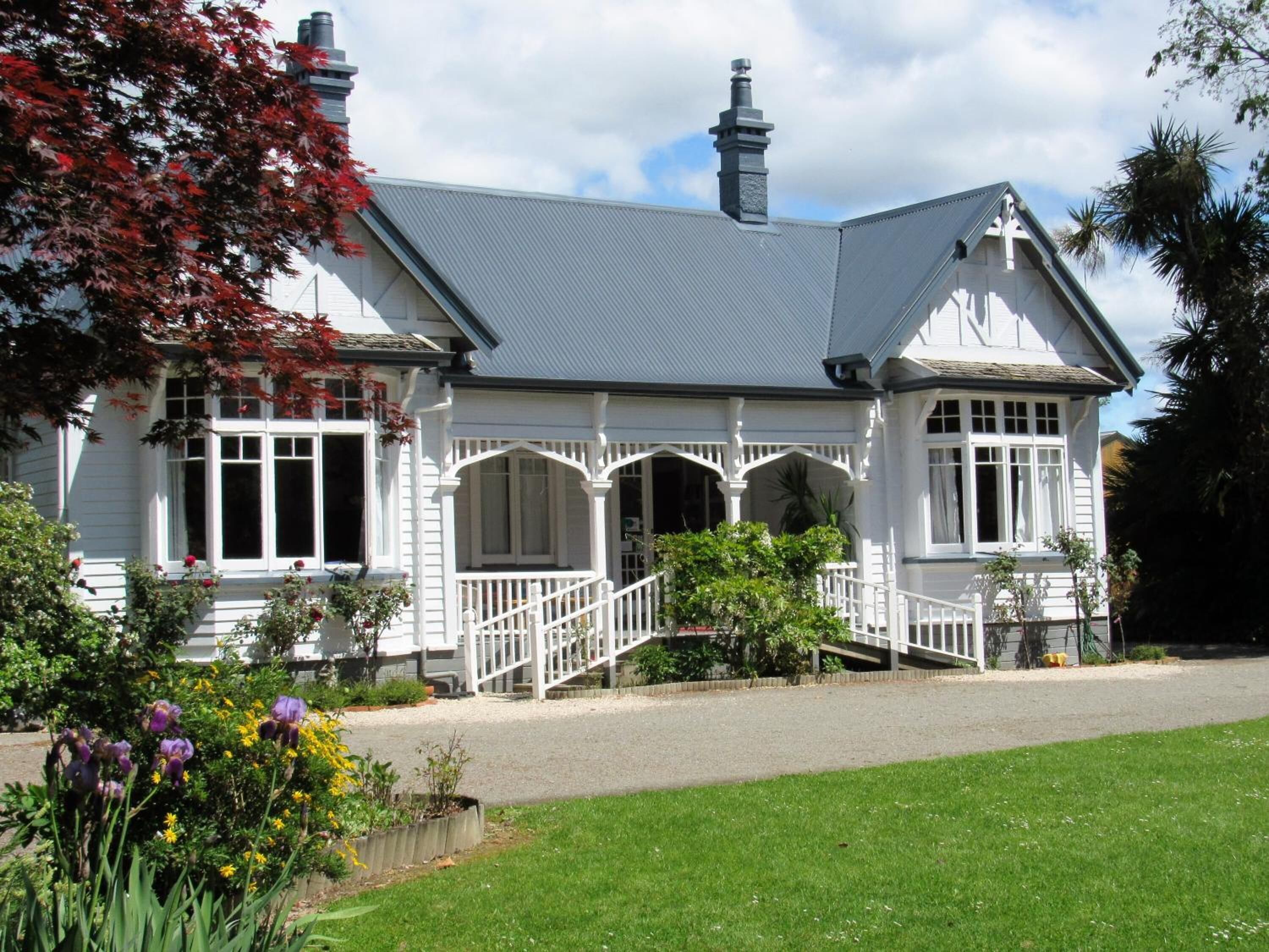 A white colonial-style house with a grey roof, a front porch with a white railing, and a small garden with flowers and shrubs.