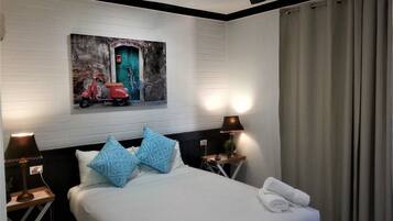 Una habitación de hotel con cama, dos lámparas de noche, un ventilador de techo, un cuadro de una scooter roja y una ventana con cortinas.