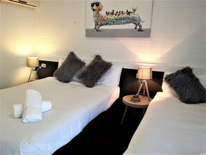 A hotel room with two beds, each with fluffy pillows and a white comforter. There is a painting on the wall above the beds, a lamp on a bedside table, and a small plant on the floor. - Kurri Motor Inn (Kurri Kurri)