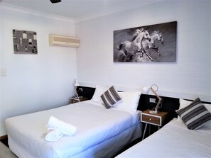 A hotel room with two beds, white bedding, a mounted air conditioner, a wall-mounted artwork of a horse, and a small table with a lamp. - Kurri Motor Inn (Kurri Kurri)