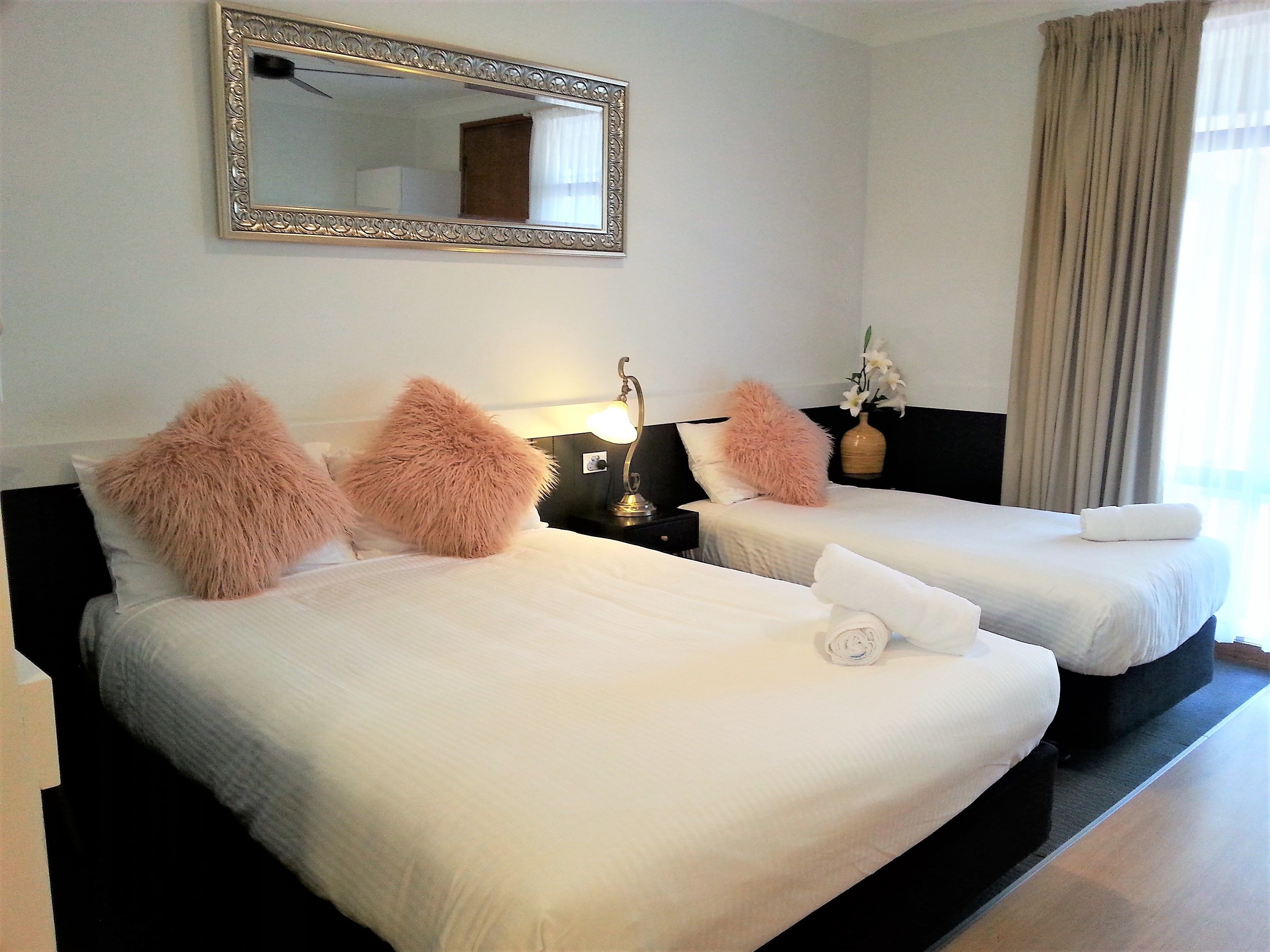 A hotel room with two beds, each with fluffy pillows, a bedside table with a lamp, and a mirror on the wall.